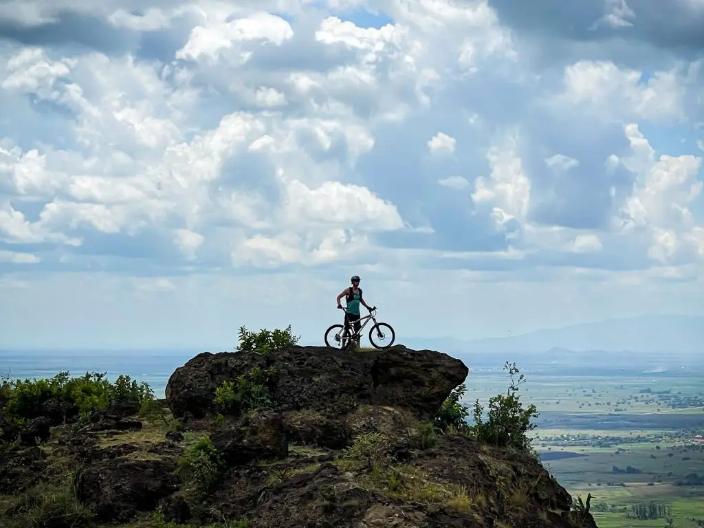 Scenic views during Sipi Biking Adventures in Uganda. Mountain Cyclist standing with a bike on a rocky viewpoint overlooking the flat plains of Mount Kadam in Karamoja region.